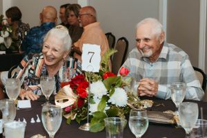 An elderly couple shares a joyful moment at a formal dinner table with floral centerpiece and table number 7.