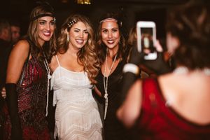 Three friends pose together at a glamorous 1920s themed party as someone takes their photo on a phone.