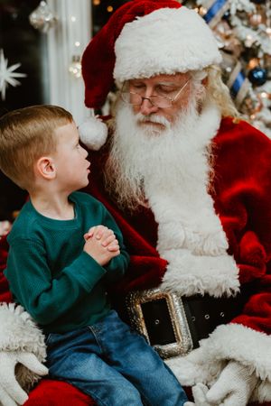 Child in green sweater sits with Santa Claus near decorated Christmas tree.
