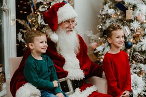 Two children in festive holiday attire sit with Santa Claus in front of a decorated Christmas tree.