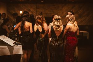 Four women in flapper-style dresses stand with their backs to camera at a vintage party.