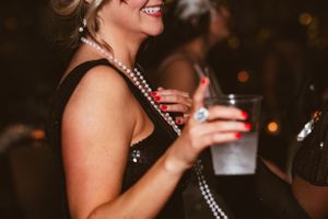 A guest in a black dress and pearl necklace smiles while holding a drink at a party.