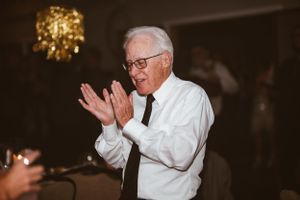 An elderly gentleman in a white shirt and tie claps enthusiastically at a celebration.