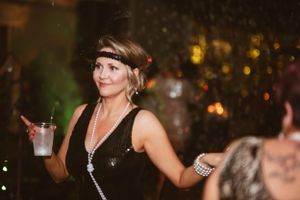 A partygoer in vintage dress and pearls holds a drink while dancing at a festive event.