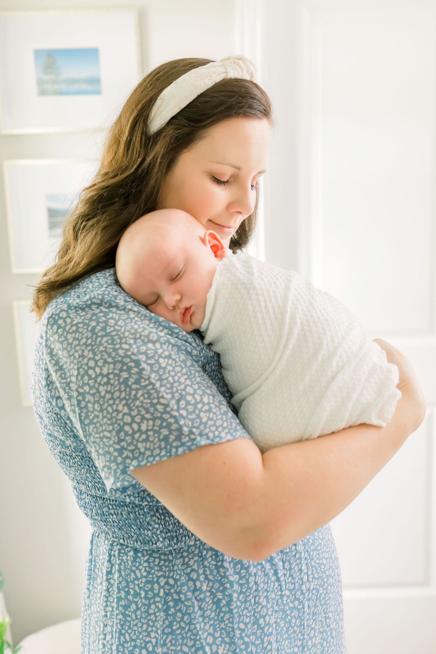 Tender moment as woman in blue floral dress cuddles sleeping infant wrapped in white.
