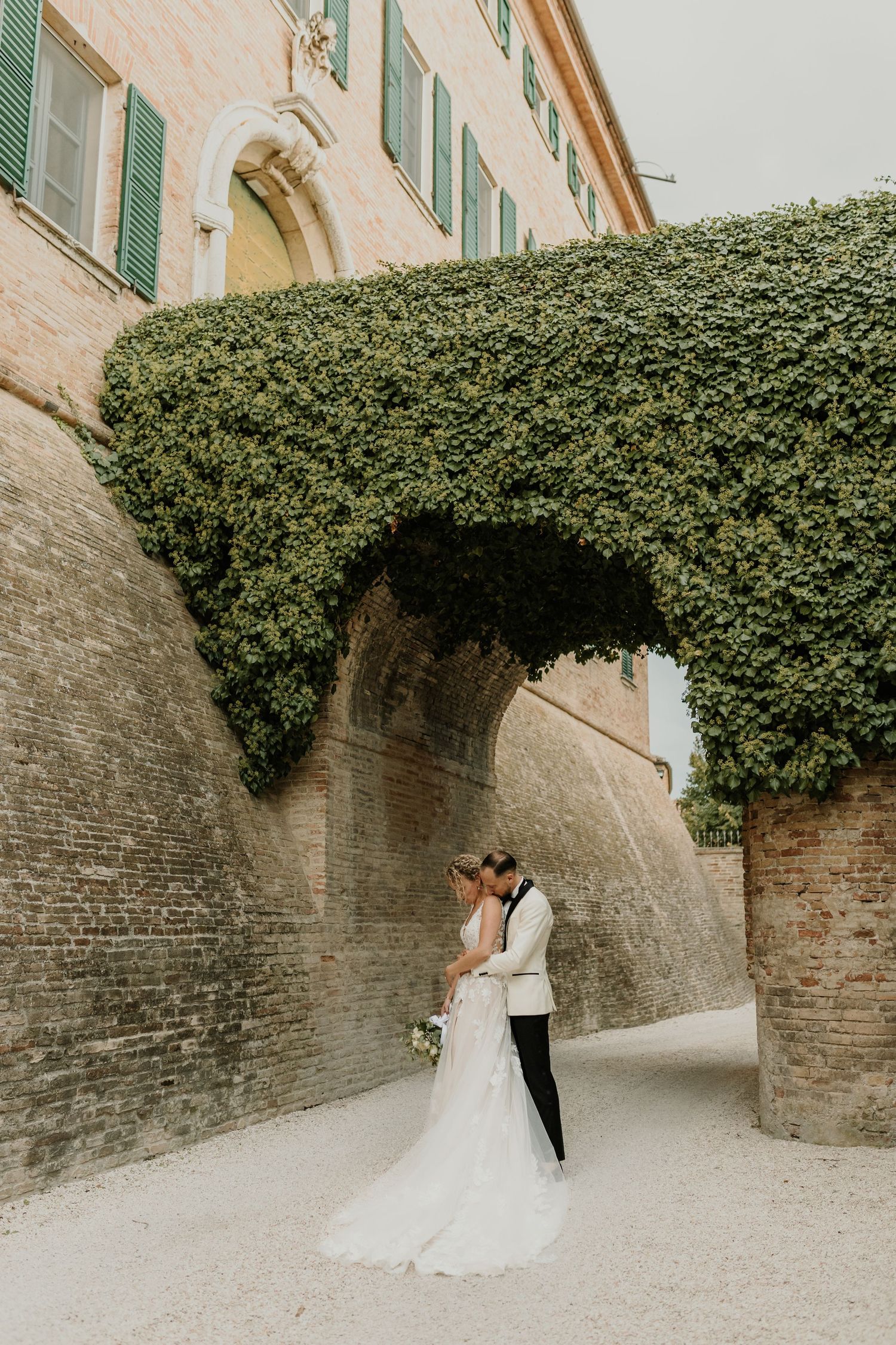A wedding couple shares an intimate moment beneath a vine-covered stone archway.