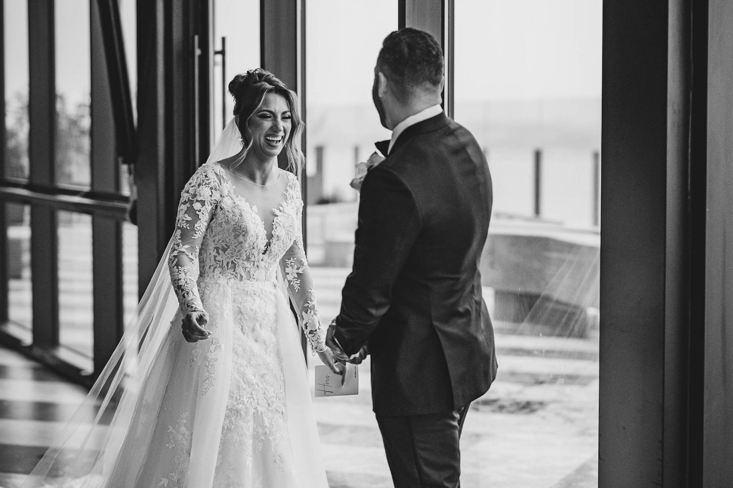 Black and white photo of newlyweds sharing a moment in front of floor-to-ceiling windows.