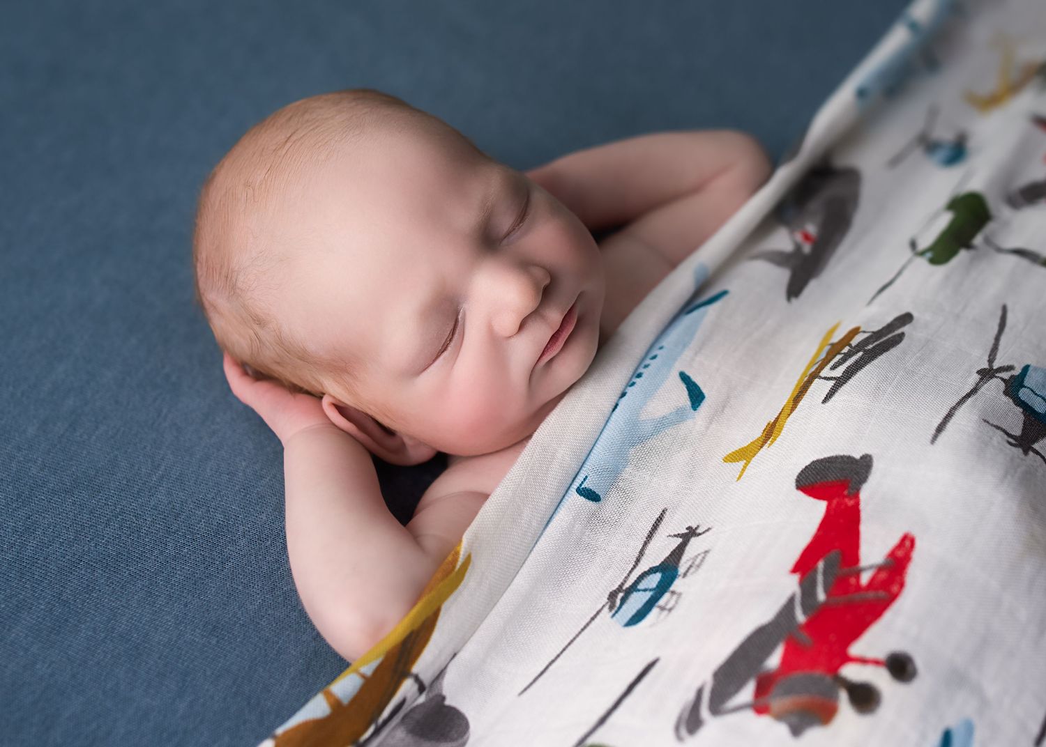 Peaceful newborn baby sleeping wrapped in a colorful muslin swaddle blanket on a blue background.