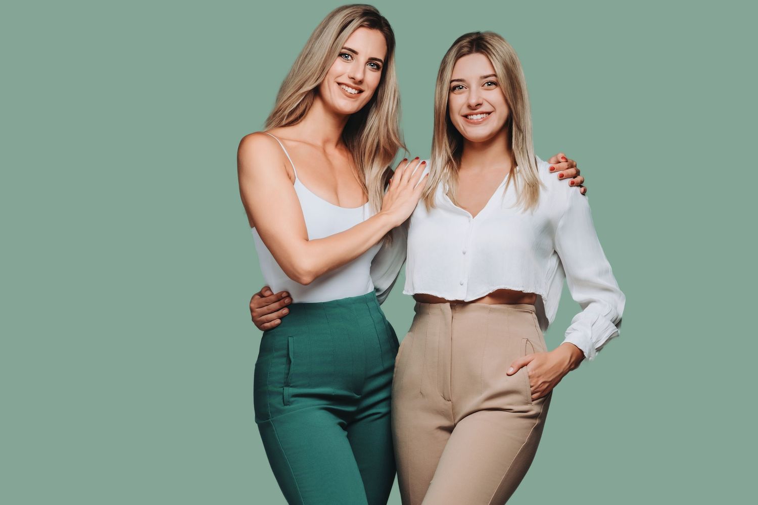 Two models pose together wearing high-waisted pants and tank tops against a sage green backdrop.