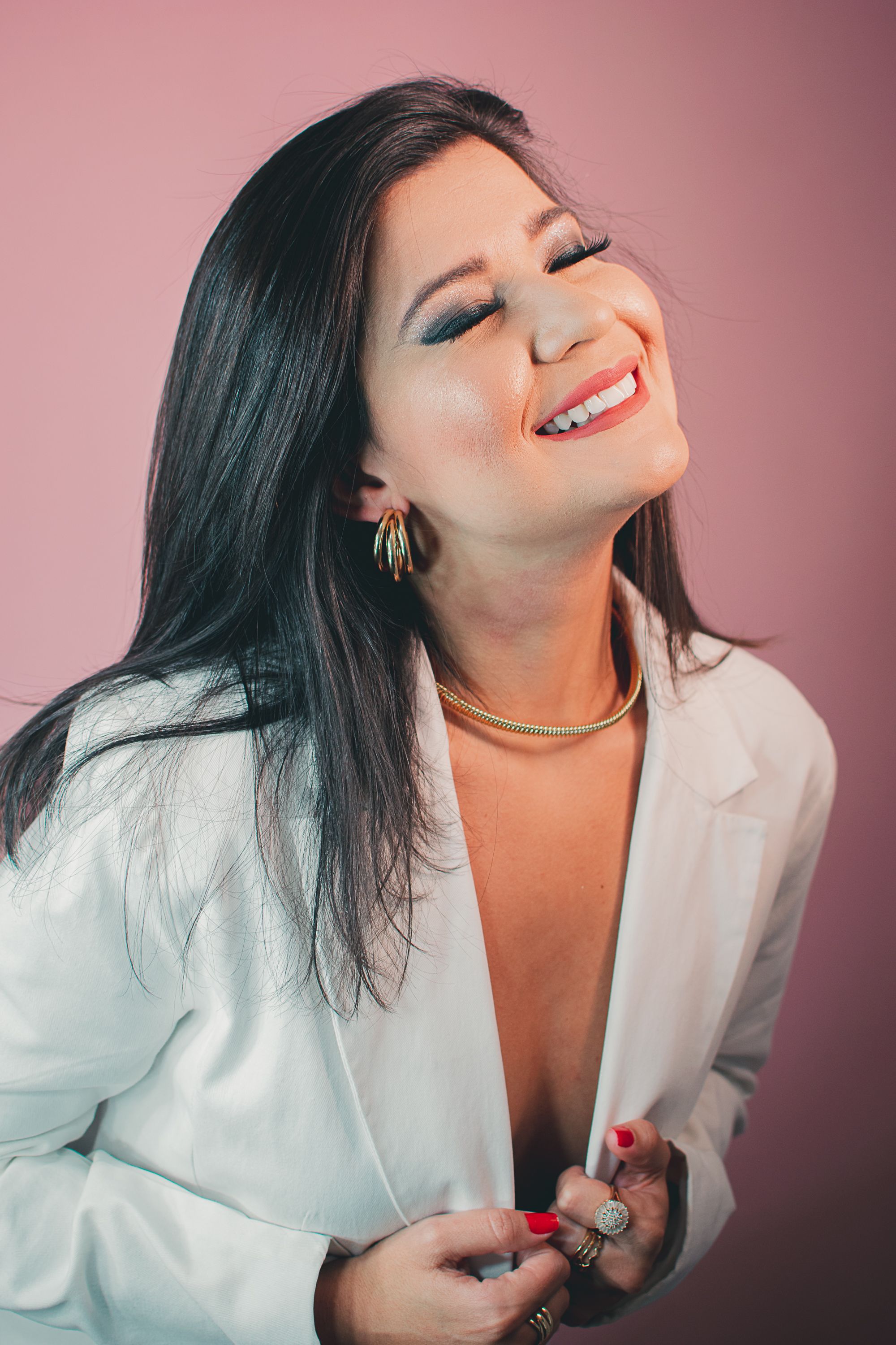 Portrait of woman in white blazer smiling against pink background with bold makeup and jewelry.