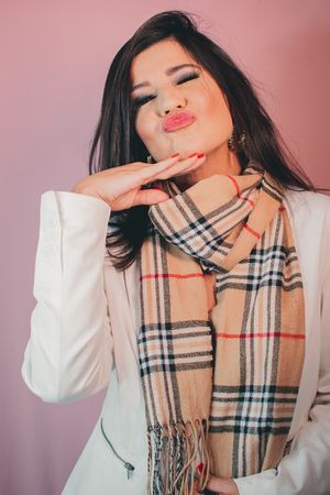 Fashion model wearing plaid scarf and white top poses playfully against pink backdrop.