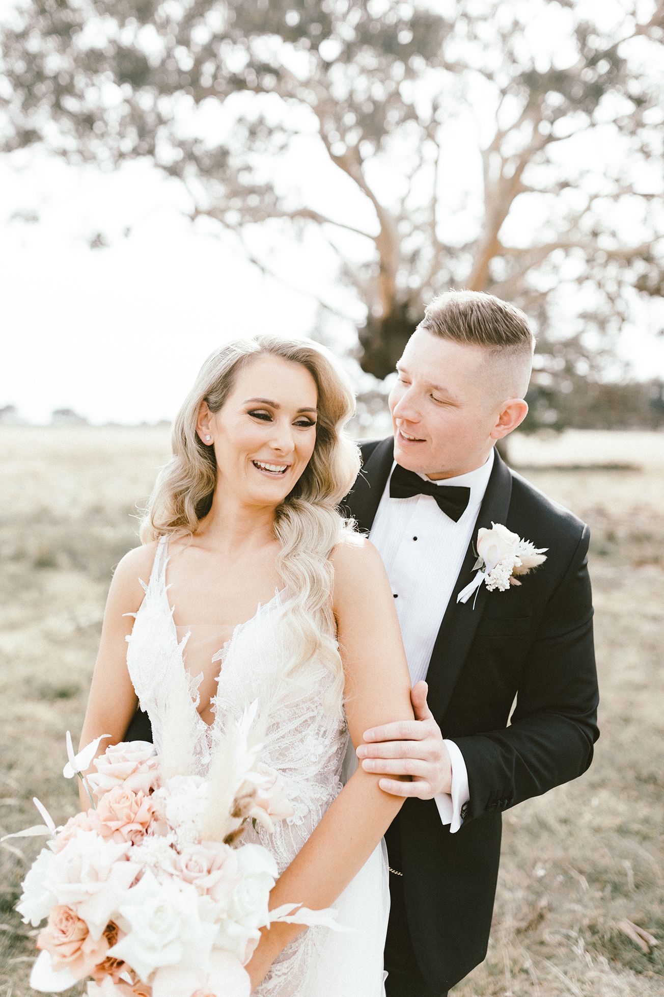 Elegant wedding couple shares intimate moment in outdoor setting with peach bouquet.