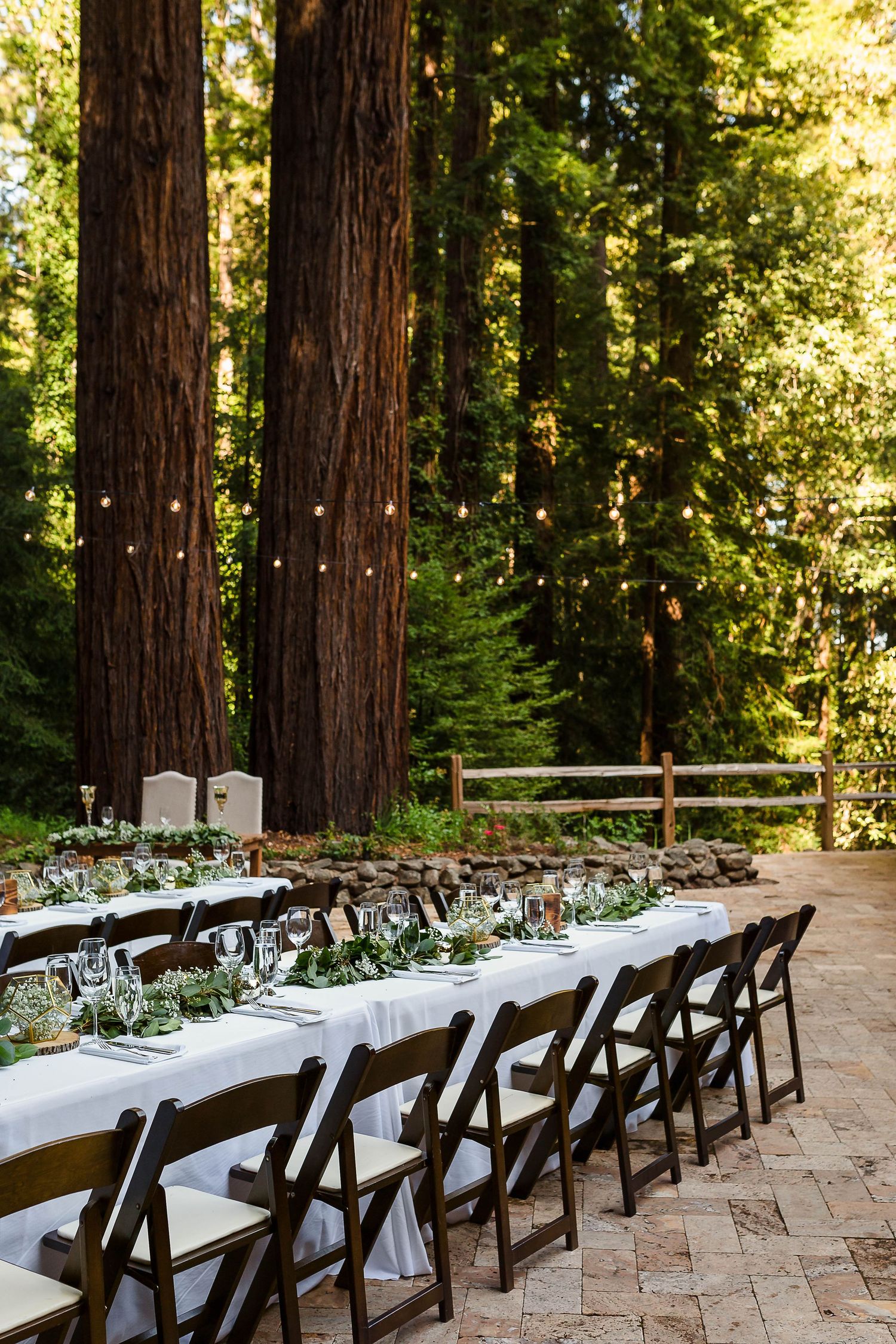 An elegant wedding reception table set among towering redwood trees with string lights overhead.