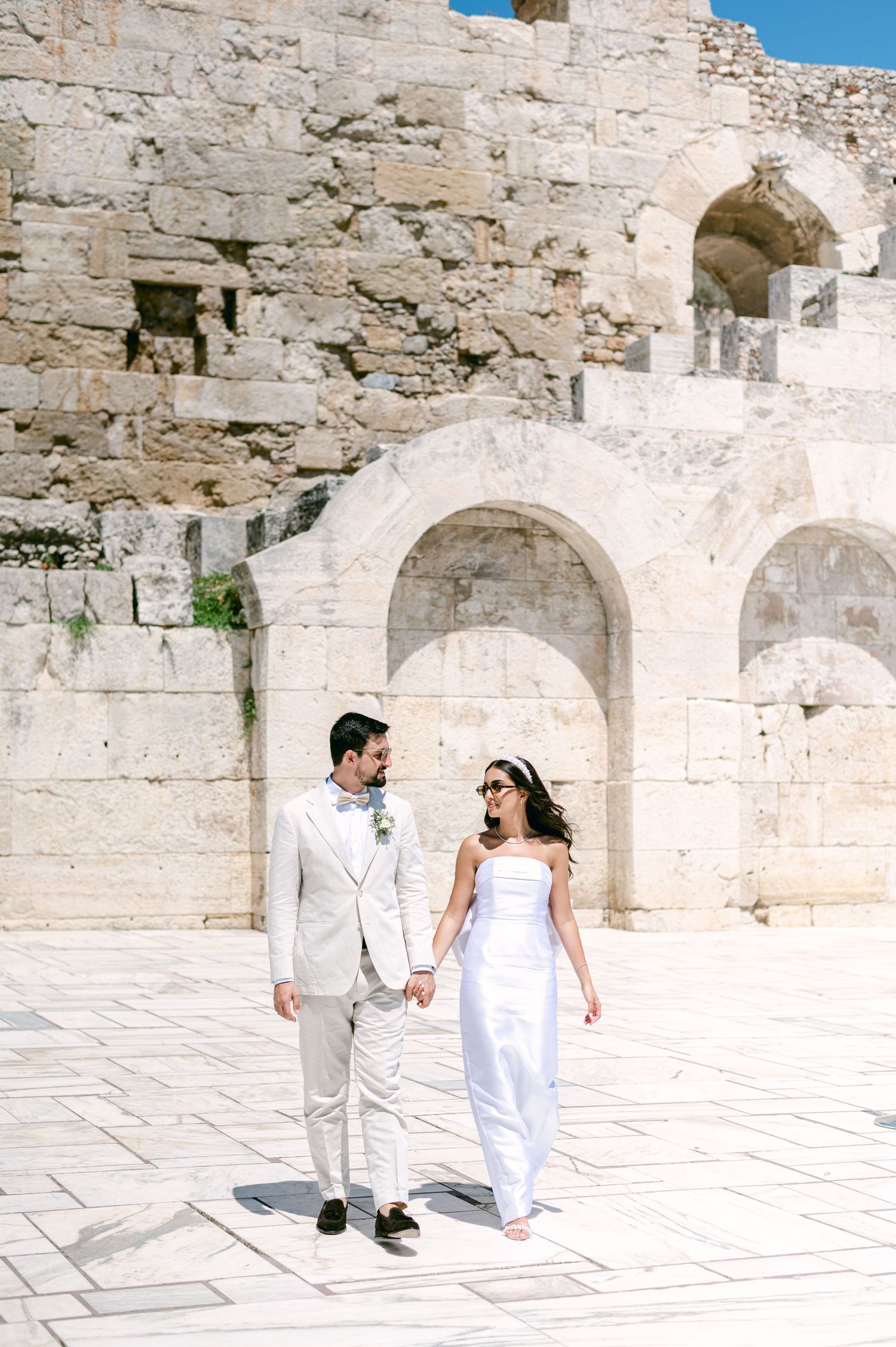 A couple in white wedding attire holds hands while walking through ancient stone ruins under blue skies.