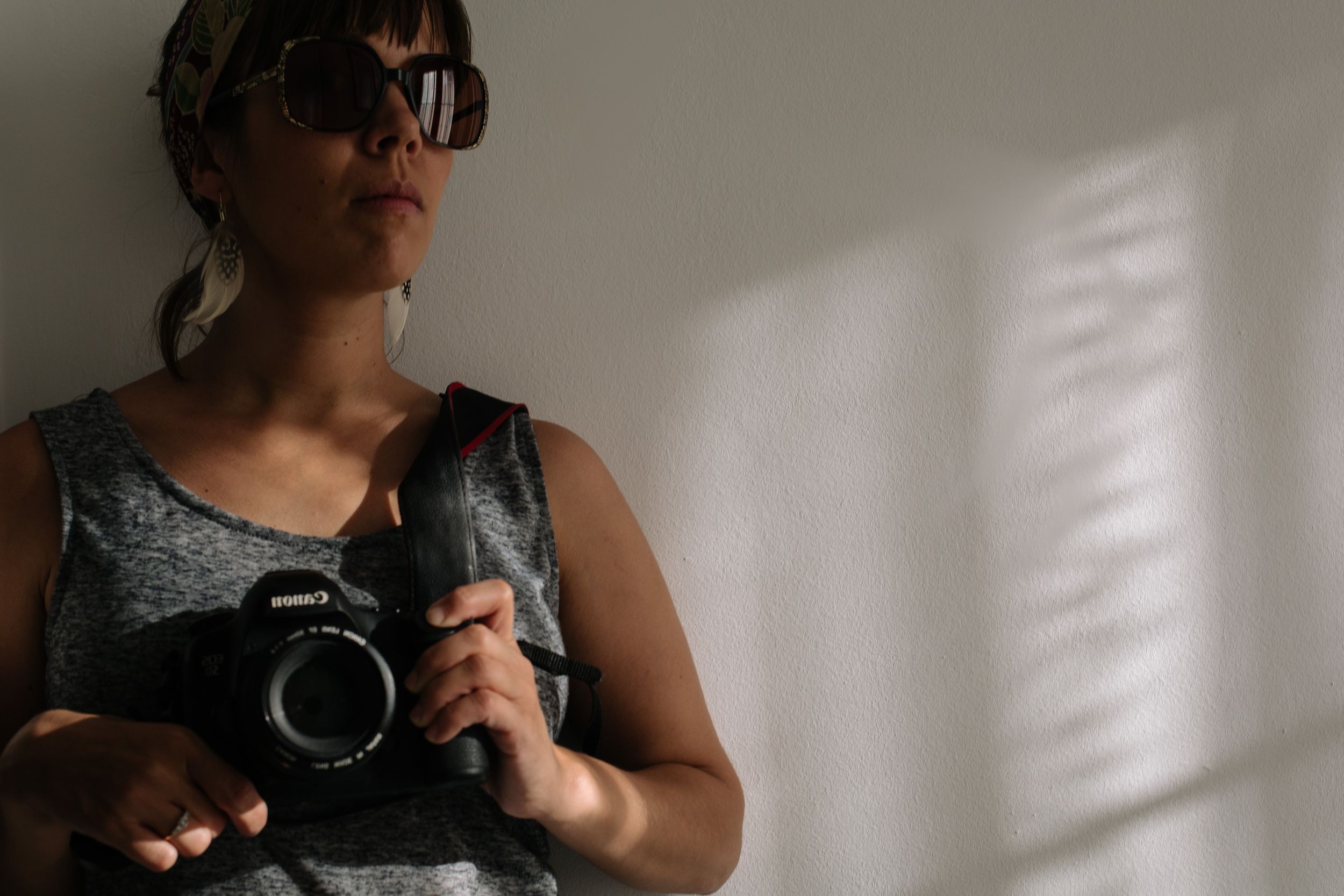 A photographer wearing sunglasses holds a camera against a white wall with window shadows.