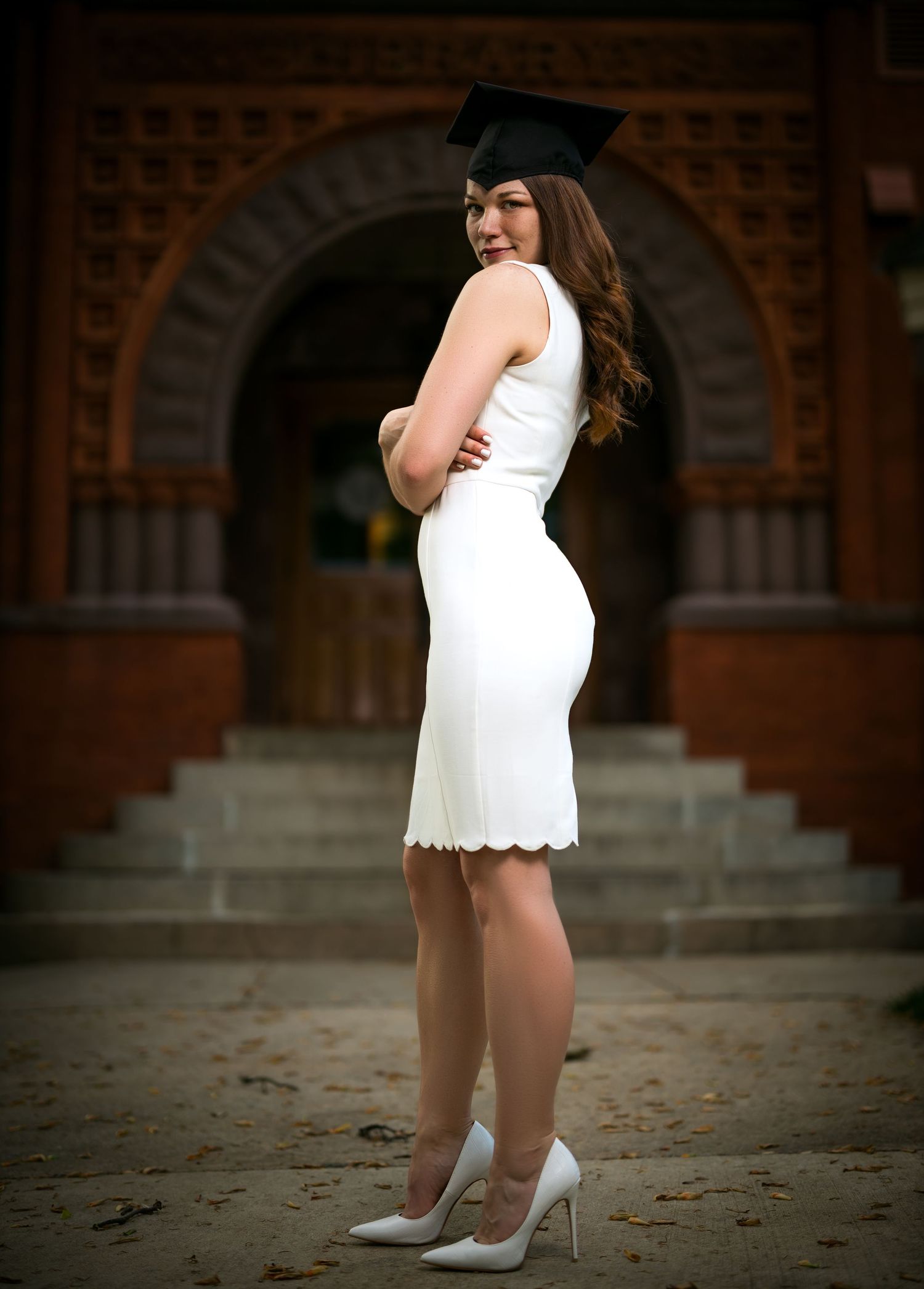 Graduate in white dress poses elegantly against brick archway and steps.