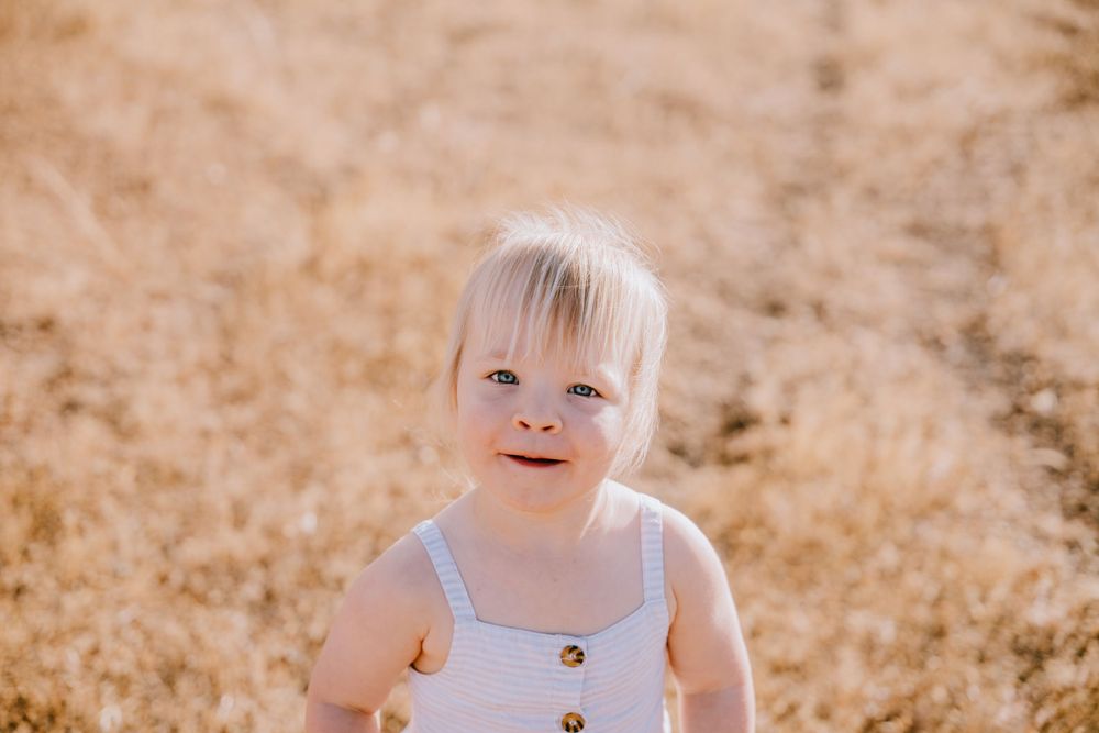 A toddler in a white outfit smiles in a sun-drenched field.