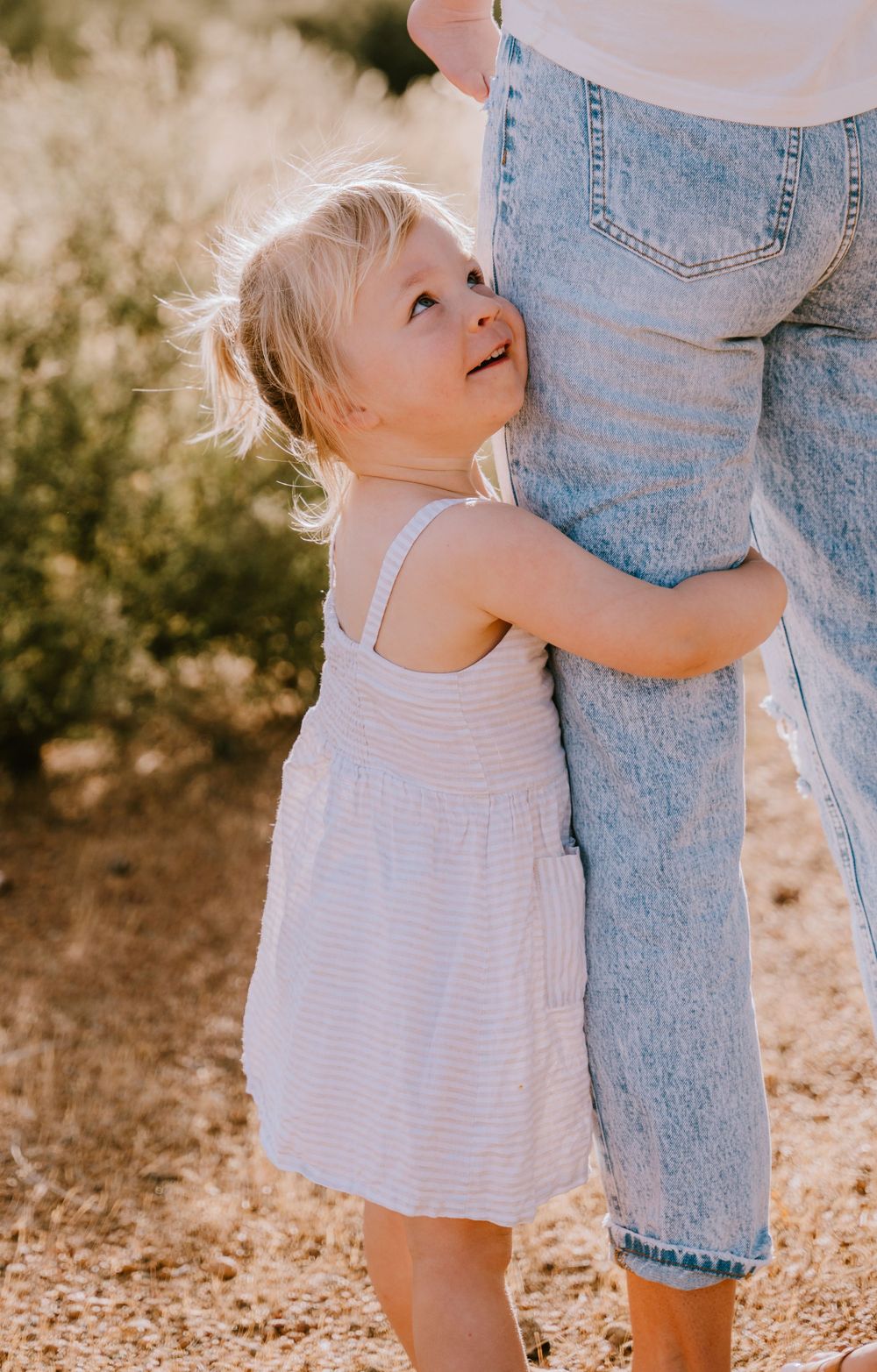 Small child in white dress hugging adult's legs in casual denim.