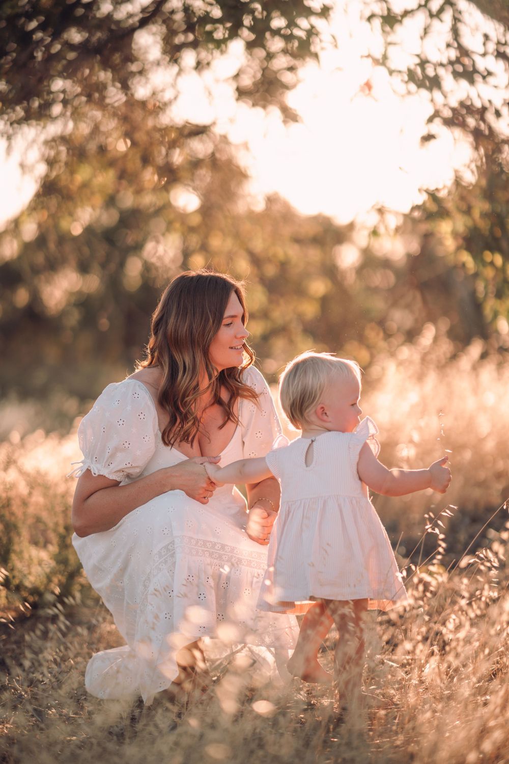 A mother and child in white dresses sit together in a sunlit field during golden hour.