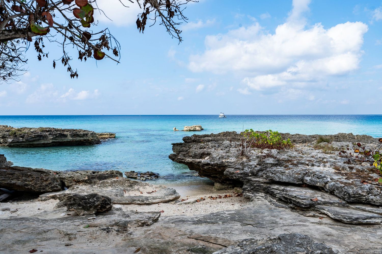 Rocky Caribbean shoreline with crystal-clear turquoise water, white sandy beach, and a sailboat on the horizon under a blue sky.
