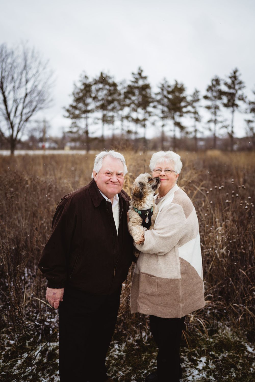 An elderly couple stands together in a winter field holding a small dog while pine trees line the background.