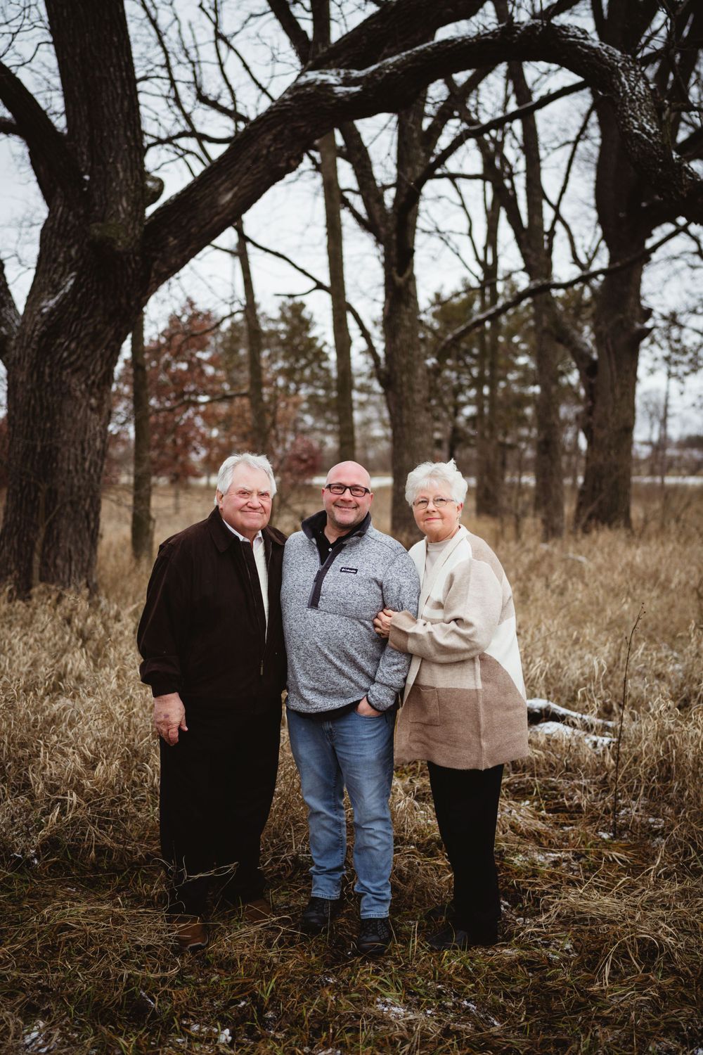 Three people stand together smiling among bare winter trees in a natural outdoor setting.