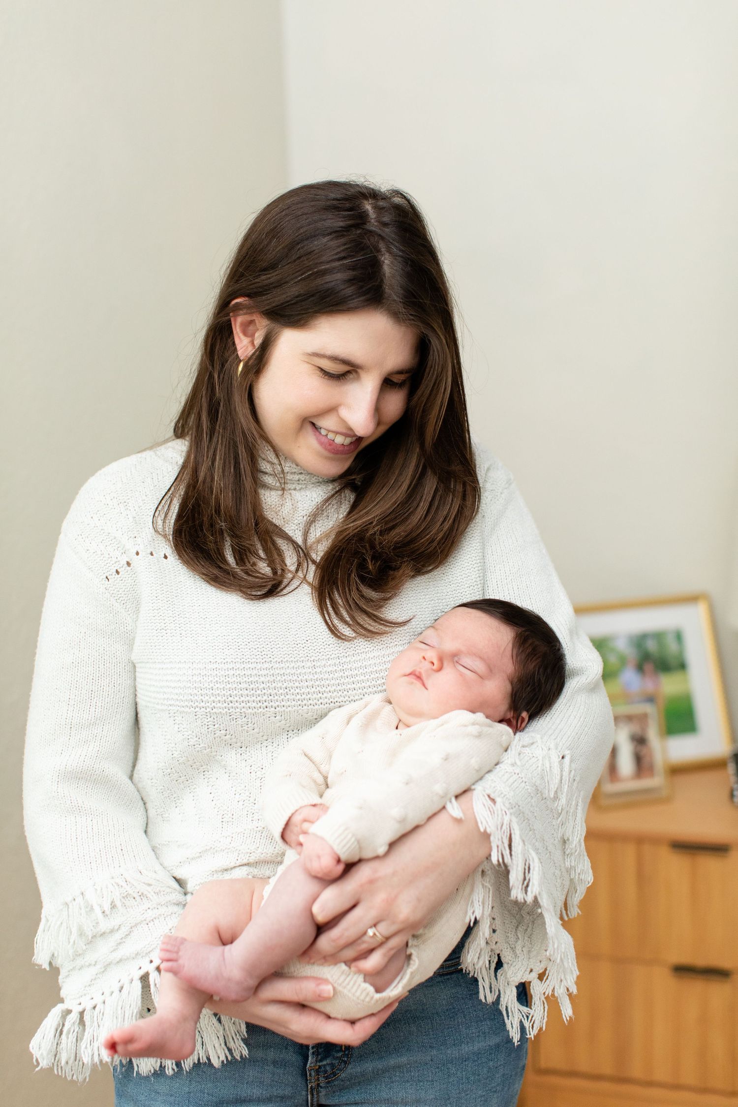 A caregiver wearing a white sweater with fringe holds and gazes lovingly at a sleeping newborn.