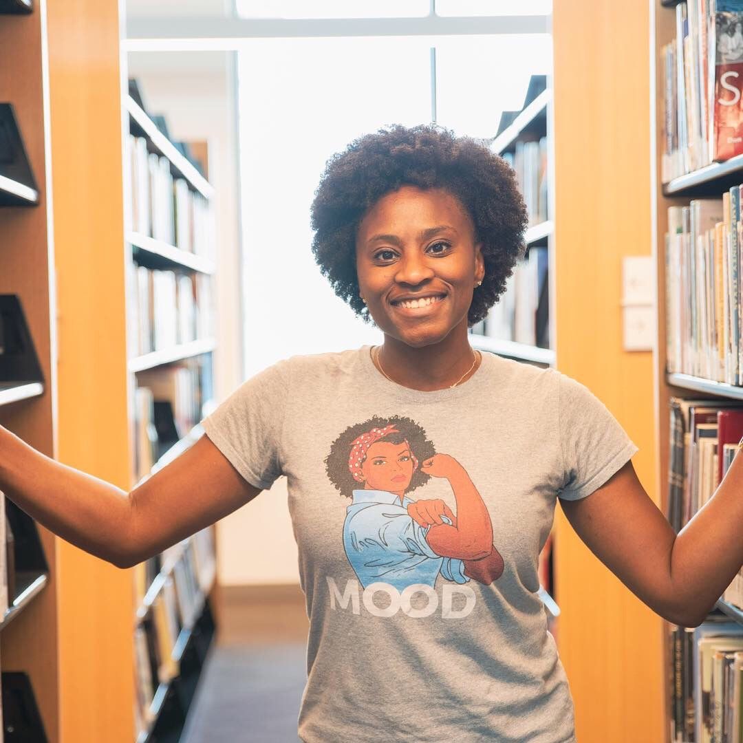 Person in beige graphic t-shirt poses cheerfully between library bookshelves.