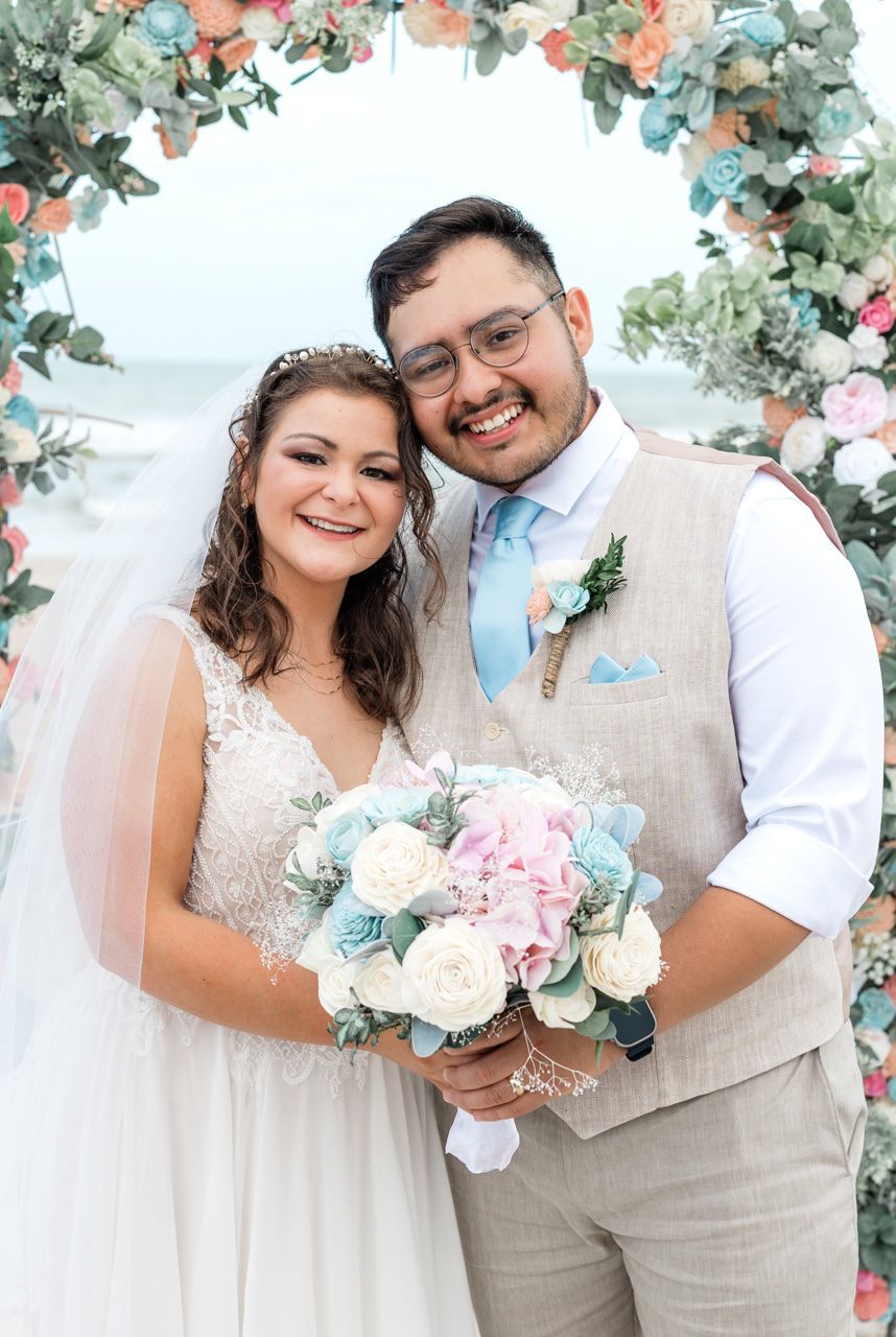 Wedding couple posing under floral arch with pink and blue flowers at beach ceremony.