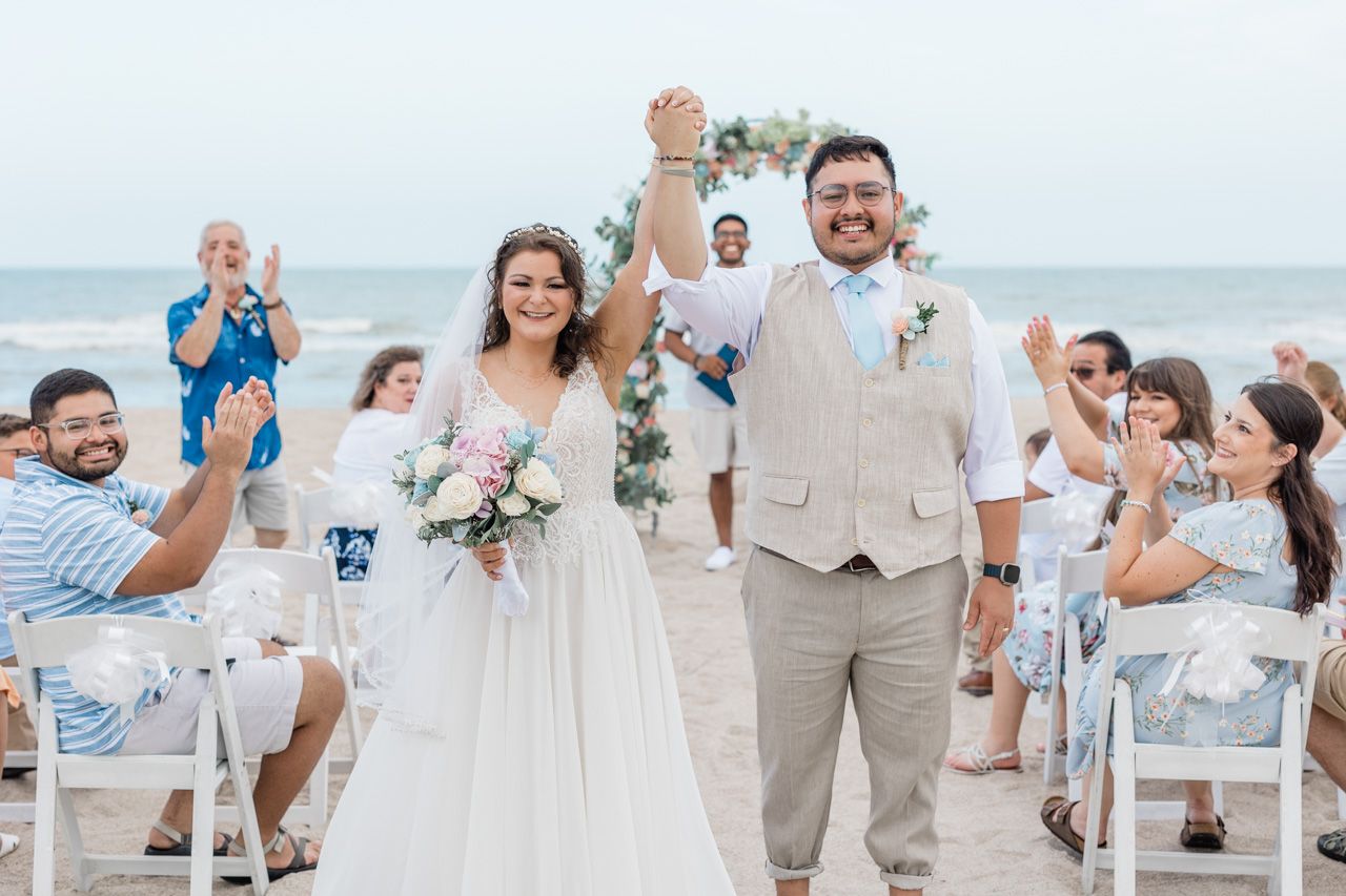 Joyful couple celebrates during beachside wedding ceremony with guests cheering in white chairs.