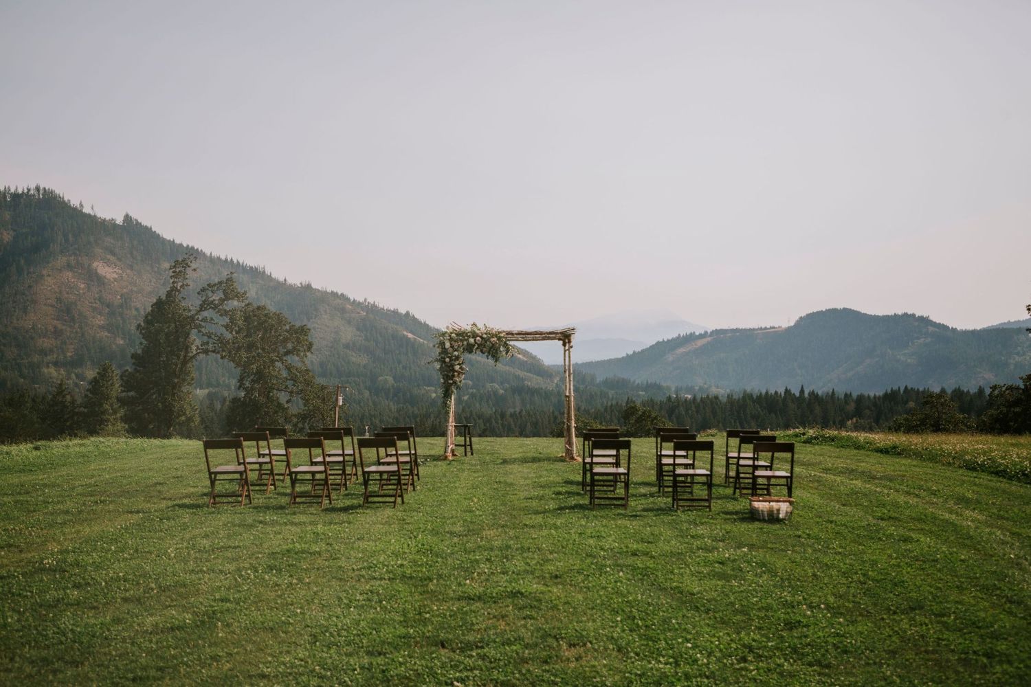 Wood chairs and arch set up for wedding ceremony in green field with mountains in the background