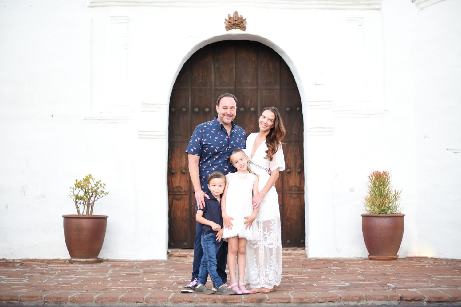 Family poses together outside historic white stucco building with arched wooden doors.