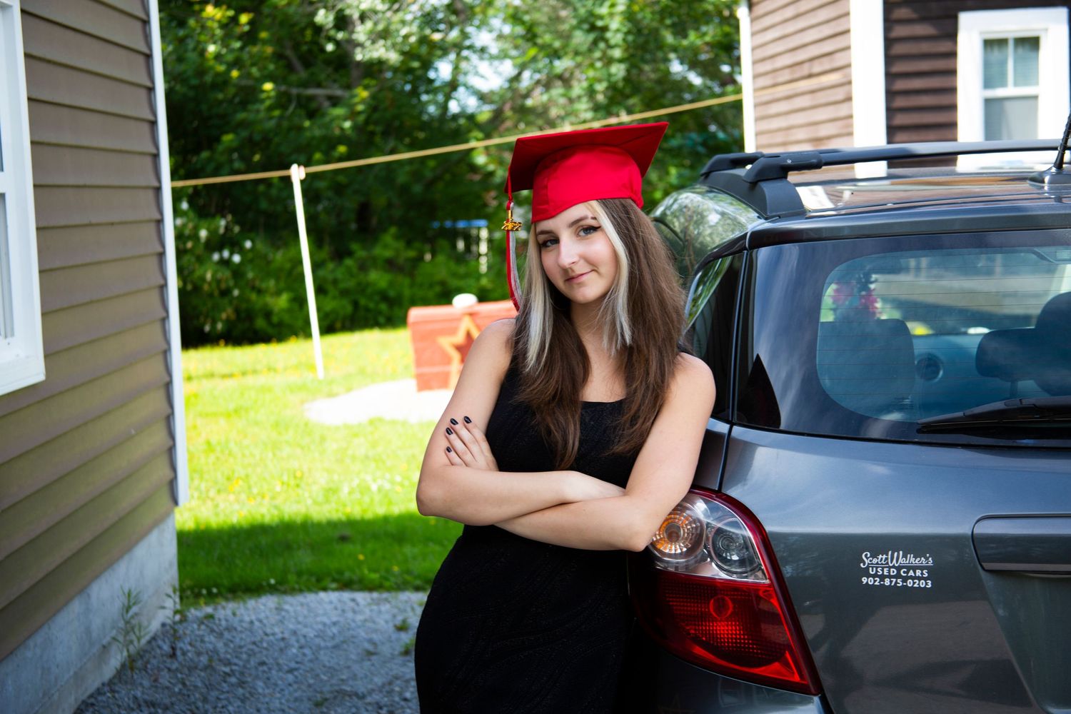 A photo of a young graduate, wearing a red graduation cap, leaning against a grey car with their arms crossed