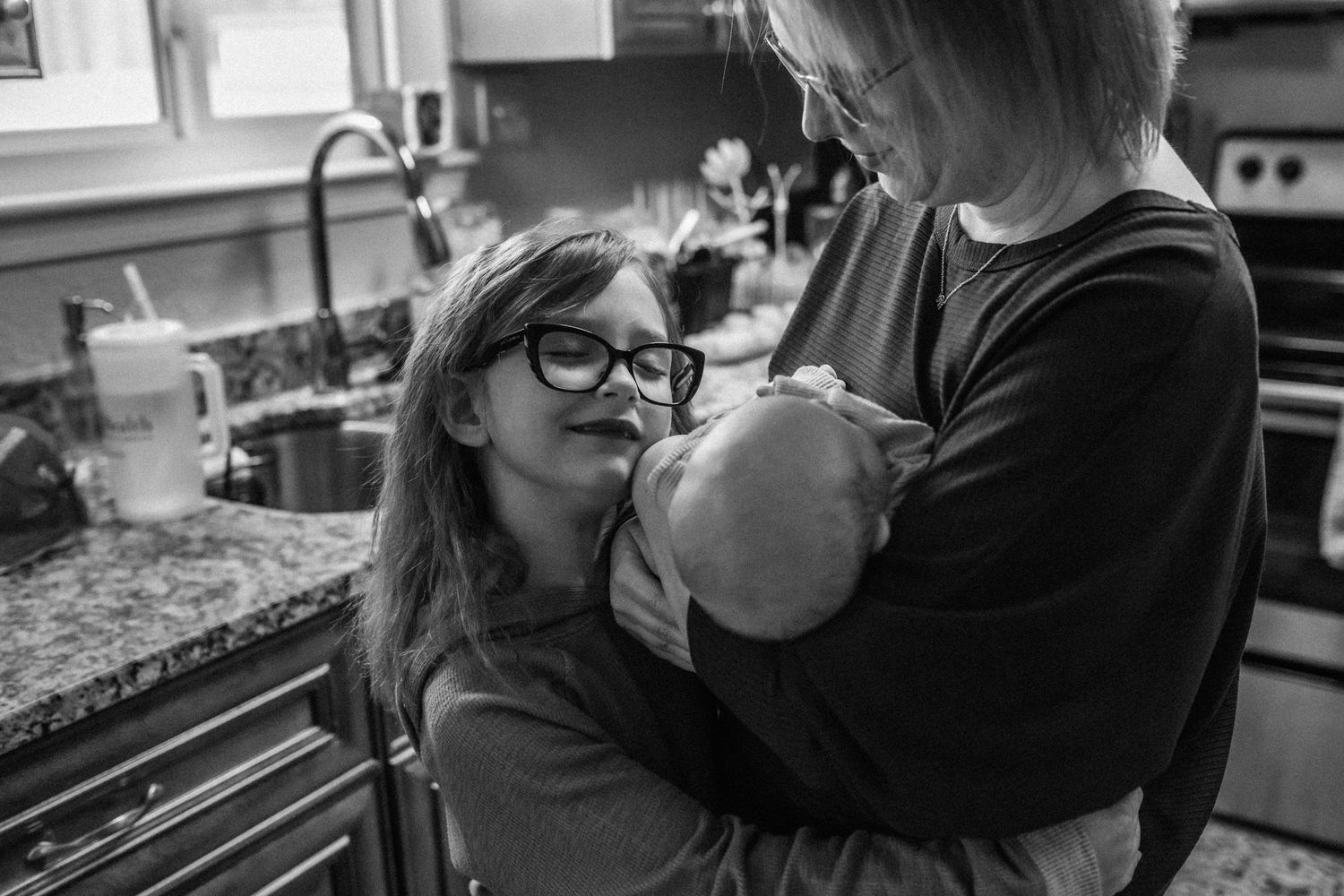 mom holding newborn baby in kitchen with big sister hugging them both in chesapeake, virginia