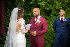 A wedding ceremony moment outdoors with bride in lace dress and groom in burgundy suit.