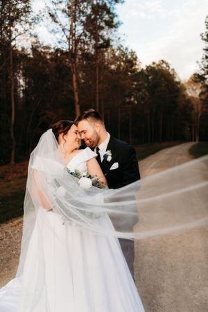 A romantic wedding photo with flowing veil captured on a wooded path at sunset.