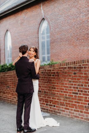 Wedding couple embraces against red brick church wall with arched windows.