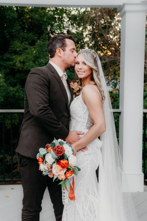 A tender wedding portrait moment on a white columned porch with coral bouquet.