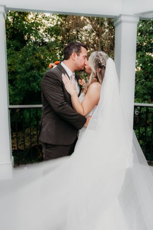 An intimate wedding kiss captured on a columned porch surrounded by greenery.