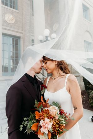Intimate wedding portrait with flowing veil and fall-colored bouquet against brick building.