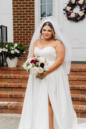 A radiant bride poses on brick steps wearing a strapless gown and holding a mixed floral bouquet.