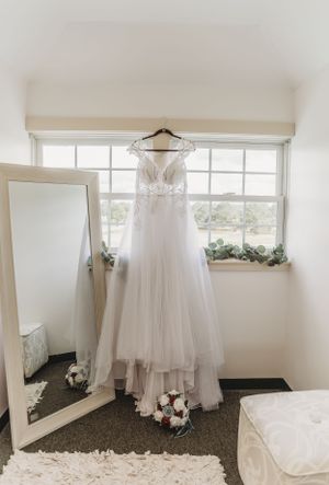 A white wedding dress with lace details hangs by a window next to a full-length mirror and bouquet of flowers.