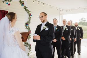 A wedding ceremony takes place indoors with a floral vine decorating the background as a wedding party stands in black suits.
