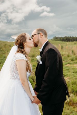 A romantic wedding moment captured in a grassy meadow with cloudy skies behind the newlyweds sharing a tender kiss.