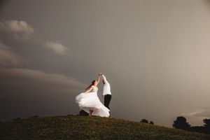Two figures in wedding attire dance atop a grassy hill at sunset with dramatic clouds in the background.