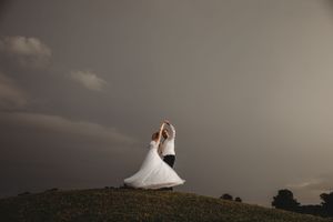 A person in a white wedding dress twirls on a hilltop against a dramatic stormy sky.
