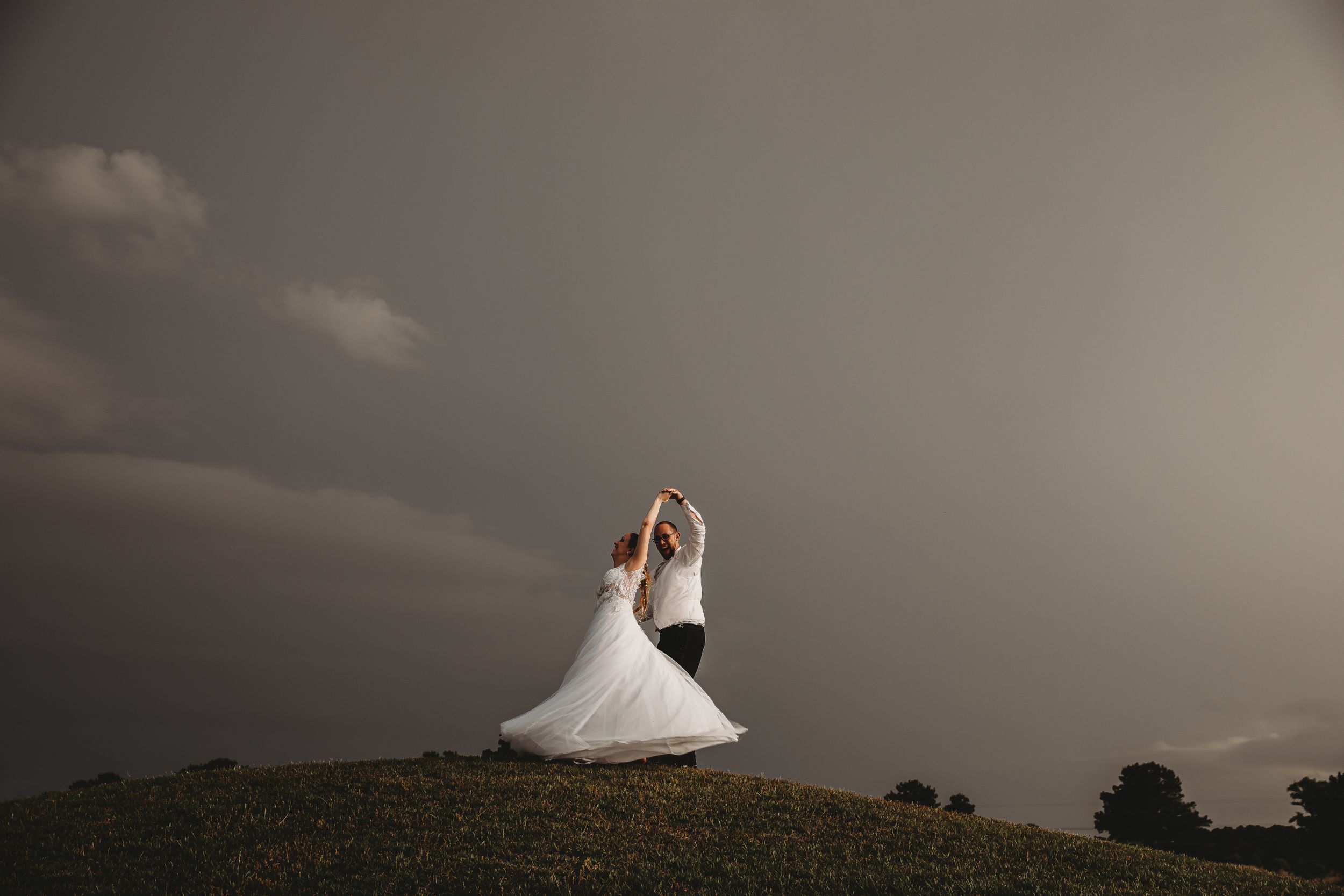 A person in a white wedding dress twirls on a hilltop against a dramatic stormy sky.