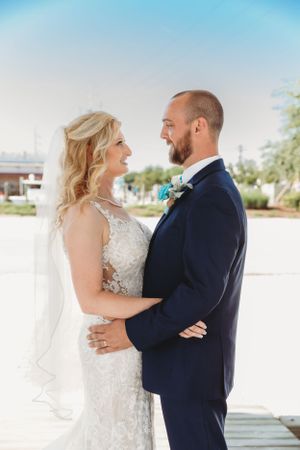 Newlywed couple embraces on a dock during a waterfront wedding, bride in lace gown and groom in navy suit.