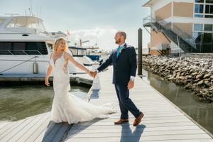 Couple walks hand in hand on a wooden dock with luxury boats and marina buildings in the background.