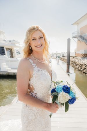 A bride in a lace wedding dress holds a bouquet of blue and white roses on a dock with boats and water in background.